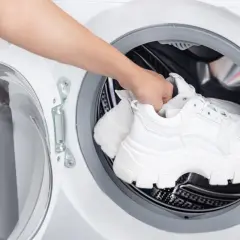 Dłoń wkłada białe sneakersy do pralki. Dowiedz się, jak prać buty w pralce, by znów były jak nowe.