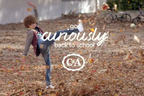 Chłopiec w jesiennych ubraniach kopie liście, z napisem "go curiously back to school" i logo C&A.