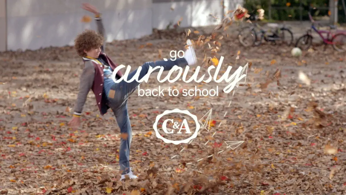 Chłopiec w jesiennych ubraniach kopie liście, z napisem "go curiously back to school" i logo C&A.